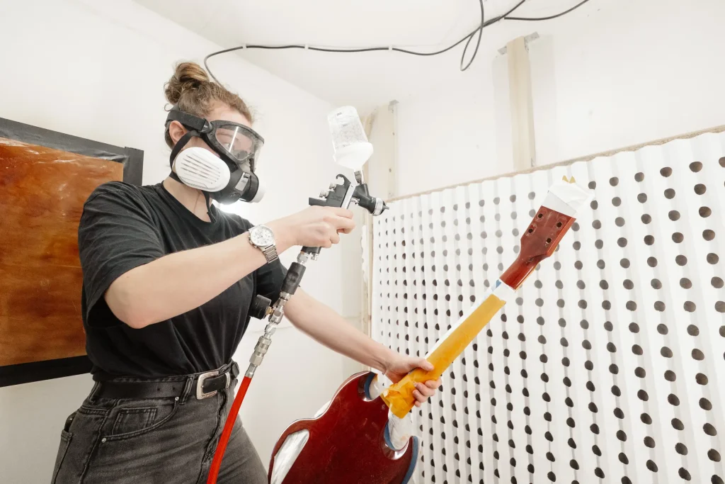 Luthier vernissant le manche d'une guitare électrique à la pistole dans une cabine de peinture